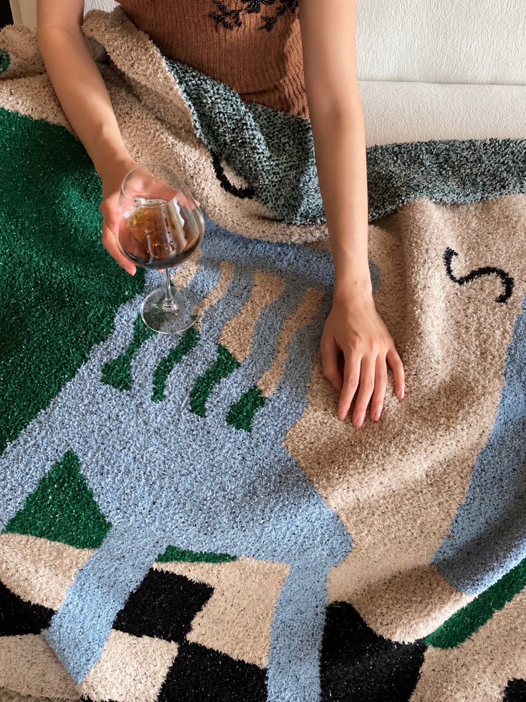 A person draped over a chunky, geometric patterned throw blanket, holding a glass of red wine. The blanket features large blocks of light blue, beige, forest green, and black.