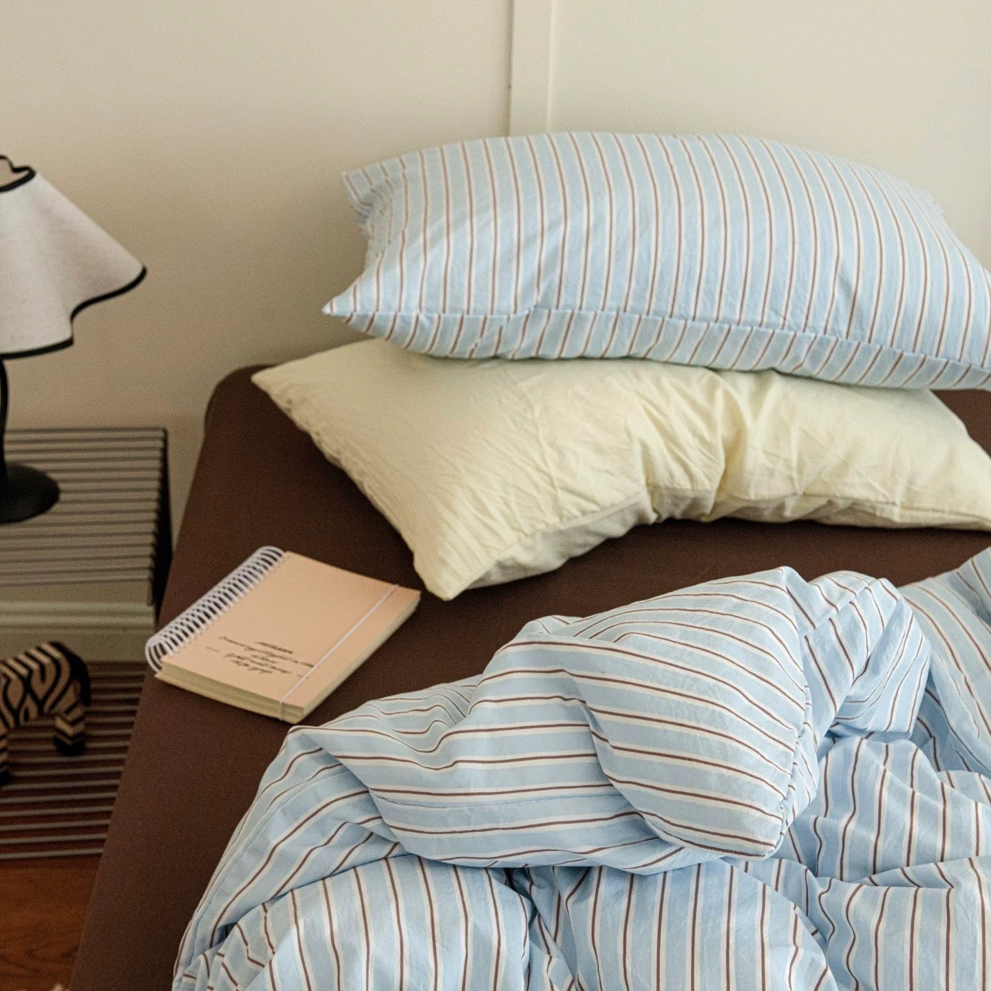 Striped bedding and pillows on a bed with a lamp and notebook in the background.