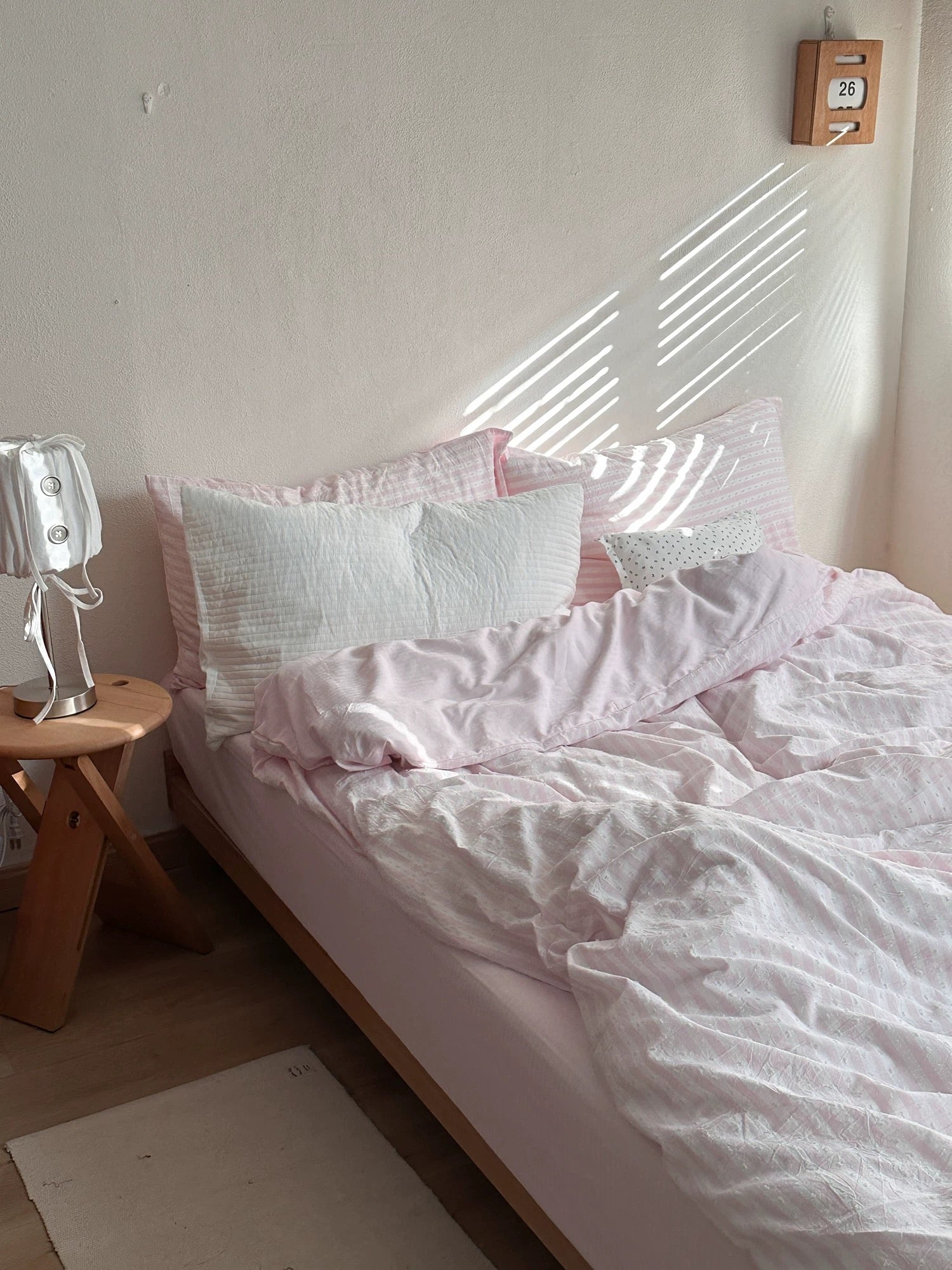 A bed dressed in light pink and white bedding, styled in a minimalist bedroom with a wooden side table, a lamp, and warm sunlight casting diagonal shadows on the wall.