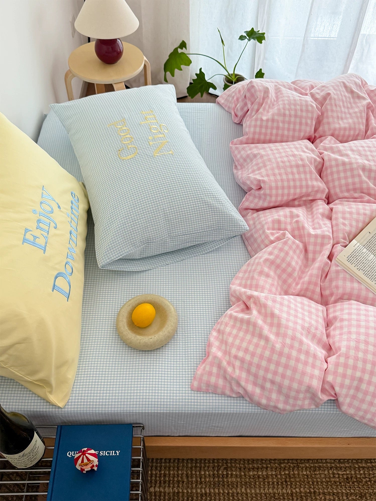 Bed with pink checkered duvet, yellow pillow, and blue pillow with text, next to a small table with a lamp and plant.