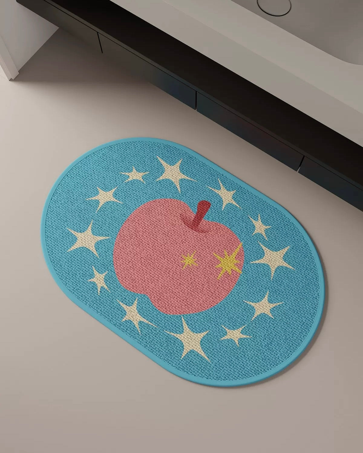 An oval-shaped blue bath mat featuring a pink apple with a yellow star and cream-colored stars, placed on a light beige floor next to a dark bathtub.