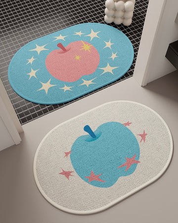 Two oval woven floor mats with apple and star designs, shown in a bathroom or entryway setting. The top mat is light blue with a pink apple and cream stars, and the bottom mat is off-white with a blue apple and pink stars.