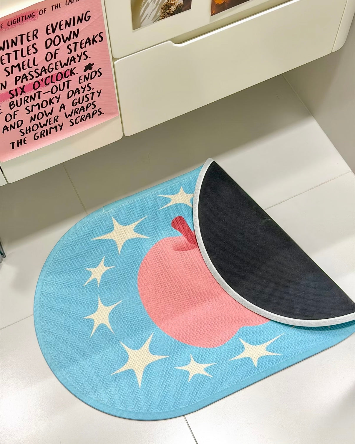 A pastel blue oval bath mat featuring a pink watermelon slice design with white star accents, partially rolled up on a white tiled floor next to a white cabinet.