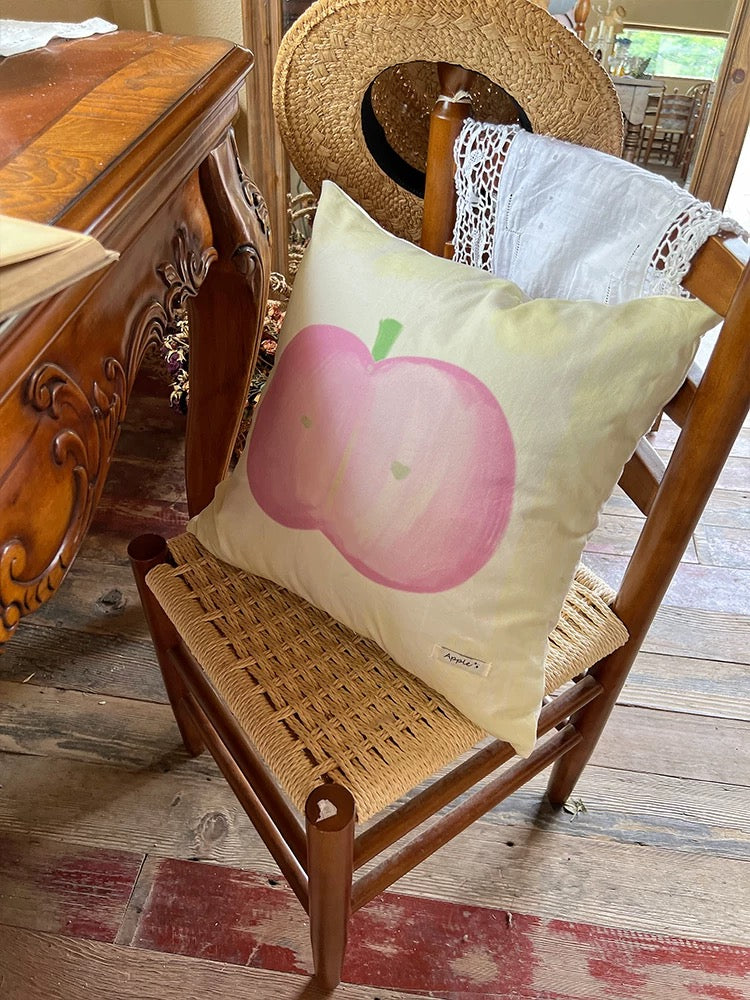 Pink apple pillow with an oil painting design on a wooden chair beside a straw hat