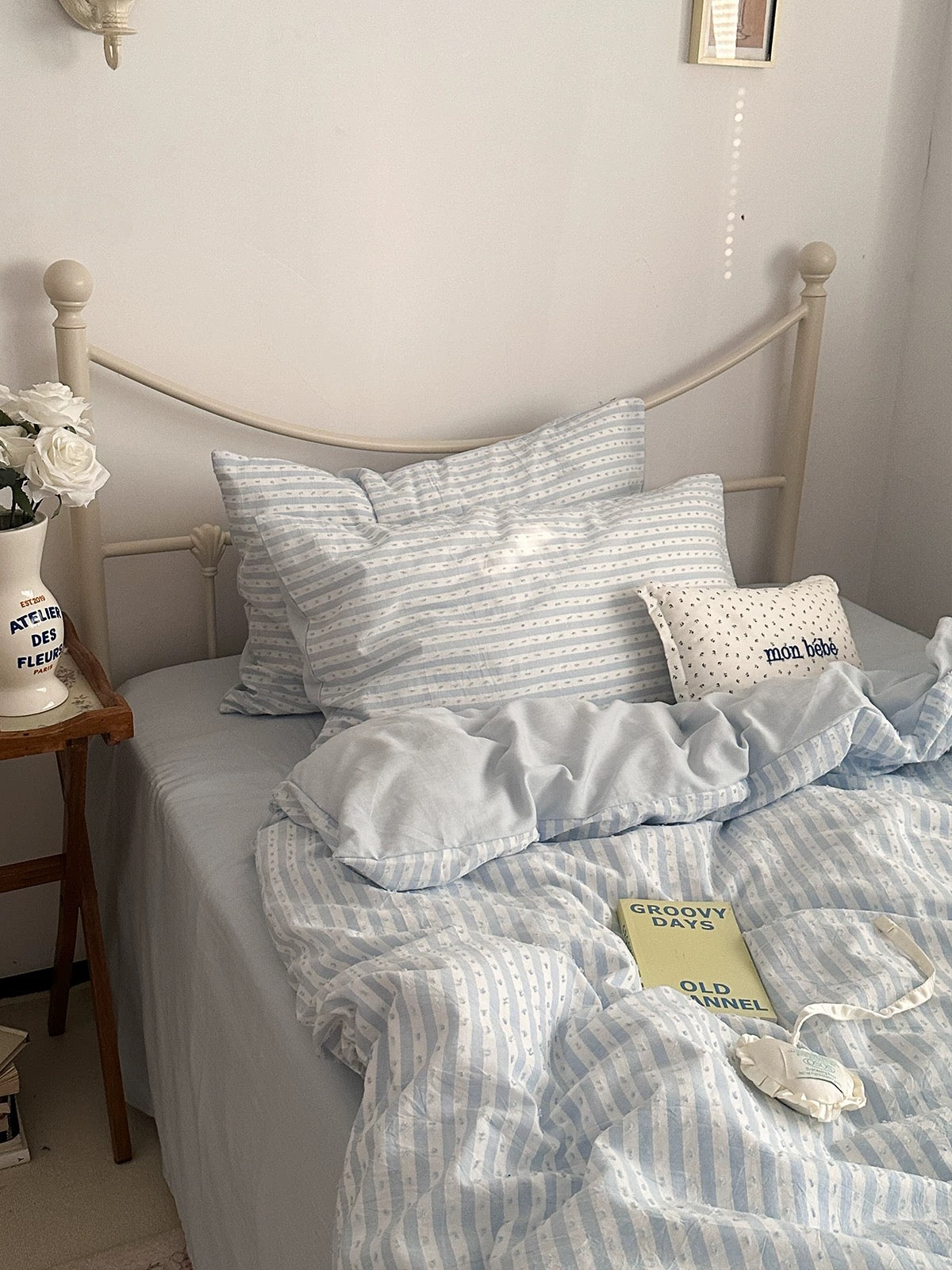 A neatly made bed in a bedroom setting featuring a light blue and white striped bedding set, including a duvet cover and two matching pillows. A small decorative pillow with white dots and the text 'mom little' is visible, along with a cream-colored metal bed frame and a bedside table with flowers.