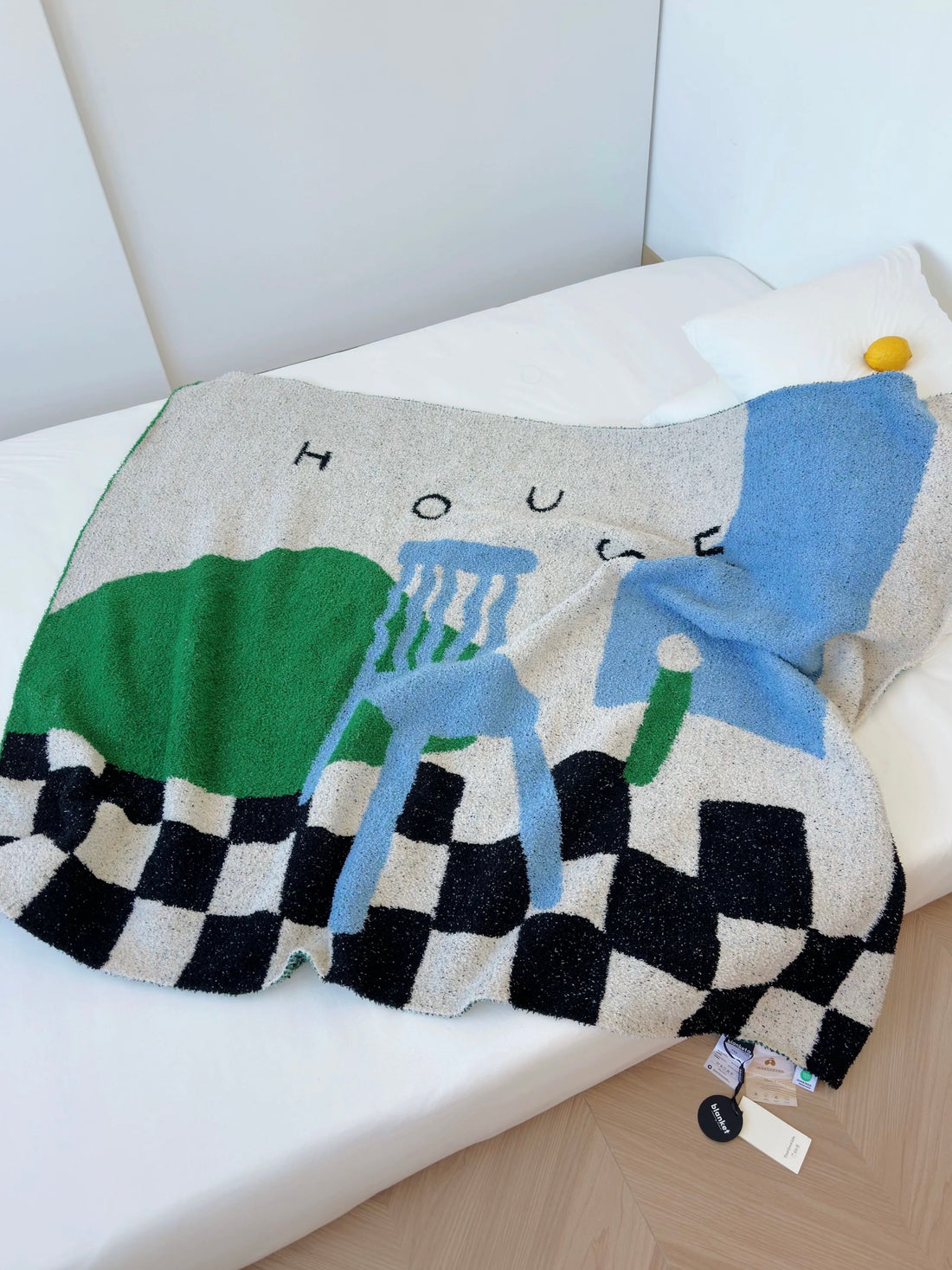 A textured knit throw blanket with a colorful abstract design and a black-and-white checkered border, draped over a white bed. The design features green, light blue, and gray blocks with the word 'HOUSE' printed above.