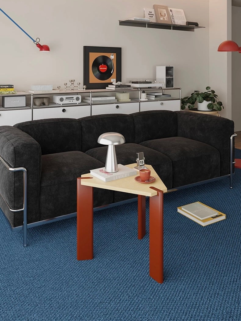 Bauhaus side table with burgundy legs and a triangular wood top holding a lamp beside a black sofa.