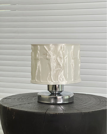Table lamp with a textured white shade on a wooden base against a window blind background