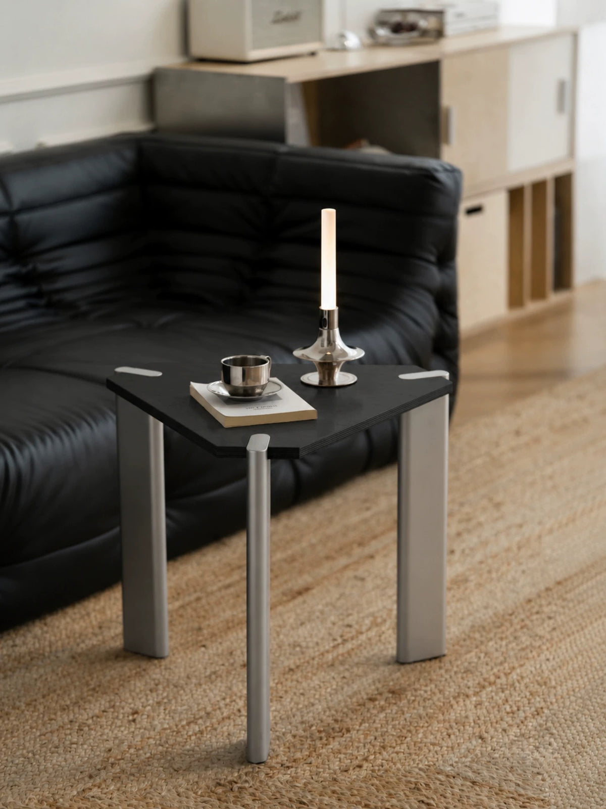 Black triangle side table with silver legs and a dark top shown beside a black sofa with a cup on top.