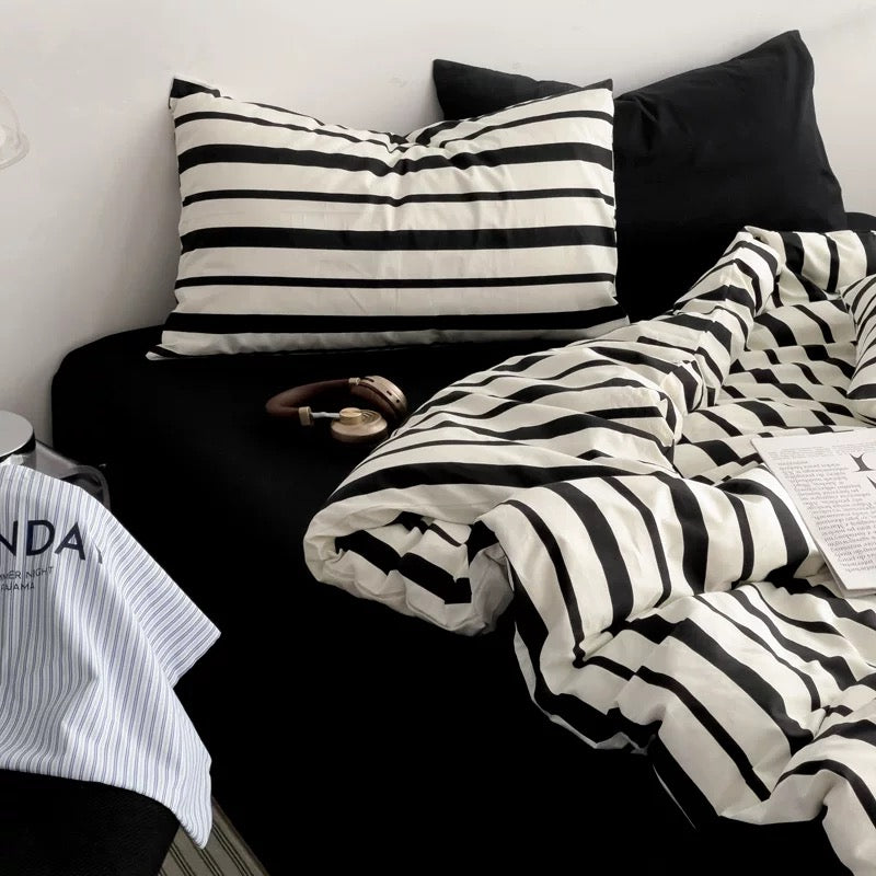 A bed styled with black and white striped bedding, including a duvet cover, pillowcases, and a folded blanket, alongside solid black pillows. A pair of brown headphones rests on the bed, with a white shirt partially visible in the foreground.