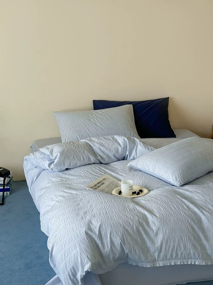 A bed dressed in light blue textured bedding with two pillows, including one dark navy pillow, set against a plain beige wall in a bedroom.