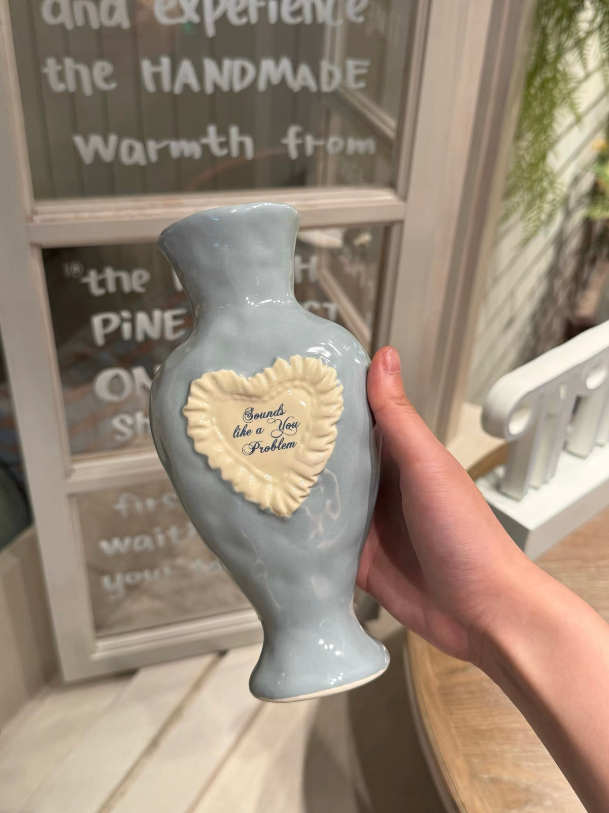 Blue ceramic vase held in one hand, showing a raised white heart, ruffled border and blue script on the front.