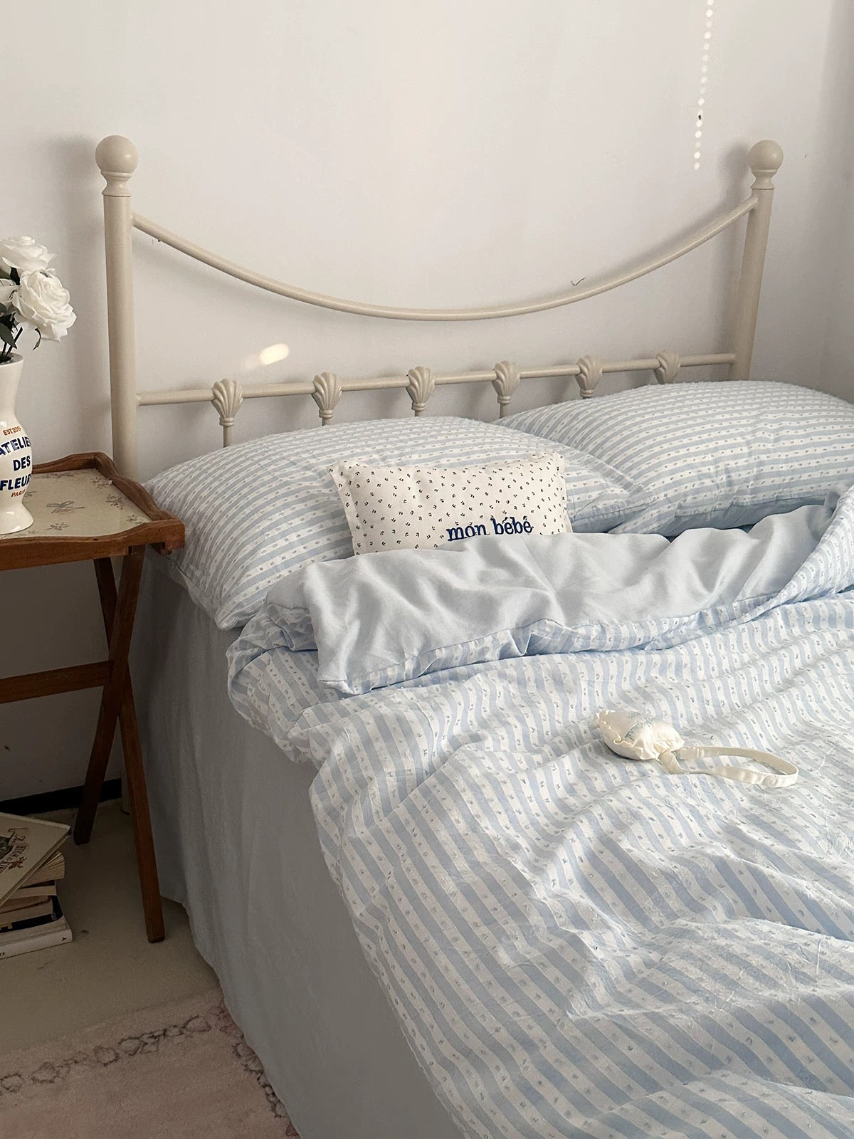 A neatly made bed with light blue and white striped bedding, including a duvet cover, pillowcases, and a small decorative pillow with the text 'mon bébé'. The bed has a cream-colored metal headboard and is situated in a bedroom next to a wooden side table.