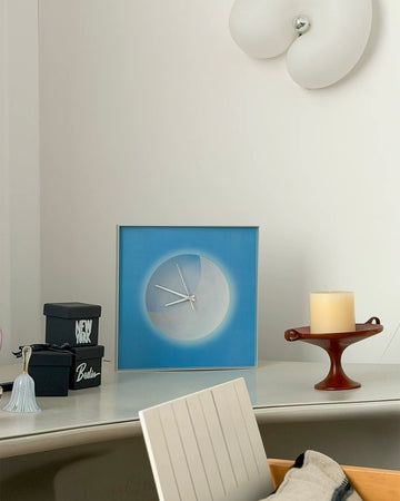 A square framed wall clock with a blue border and a minimalist white circular face sits on a light gray console table in a modern room setting, styled with a candle, decorative boxes, and a chair.