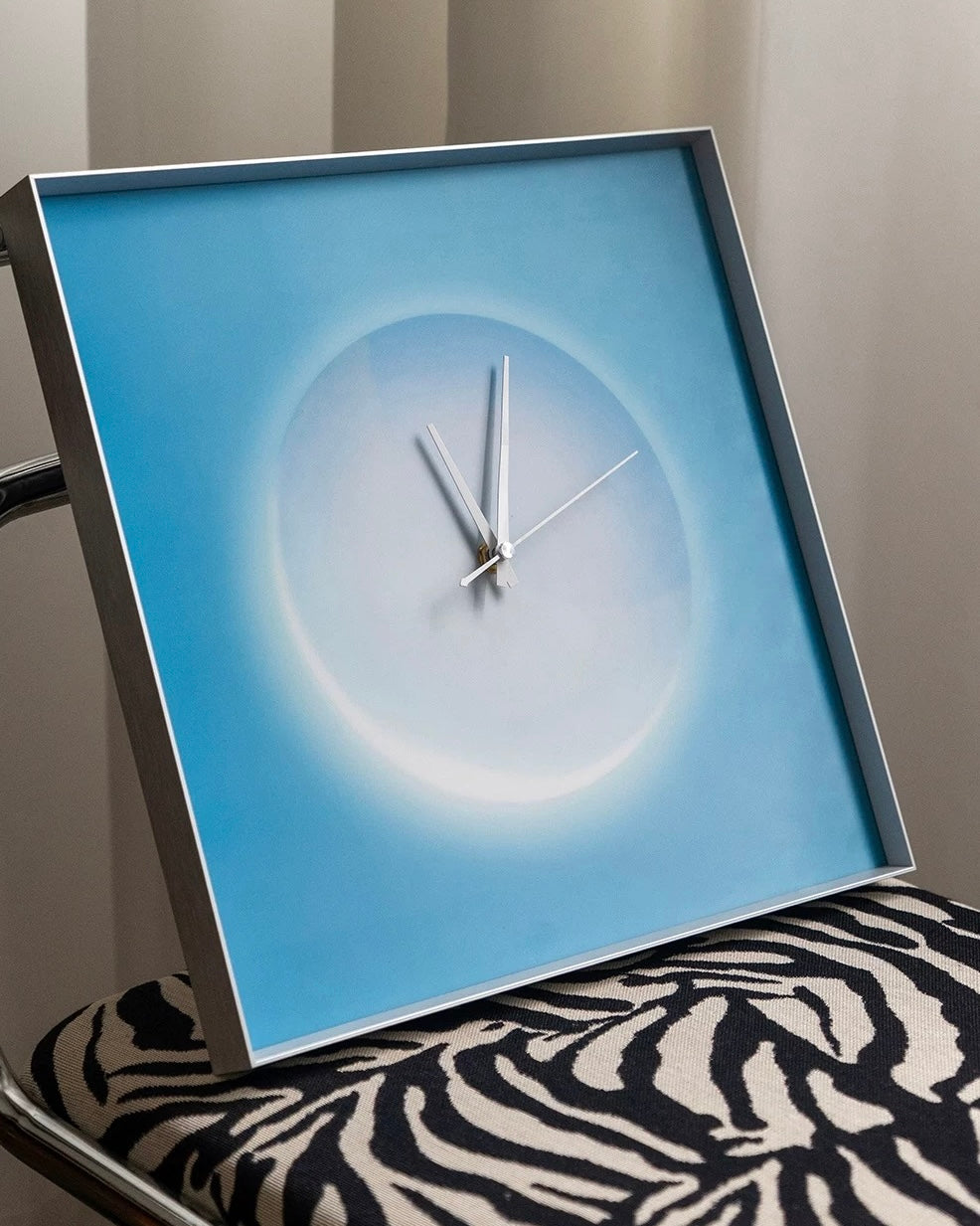 A square modern wall clock with a silver frame and minimalist white hands, featuring a light blue and white circular design on the face. The clock is shown resting on a black and white zebra-print cushion in an indoor setting.