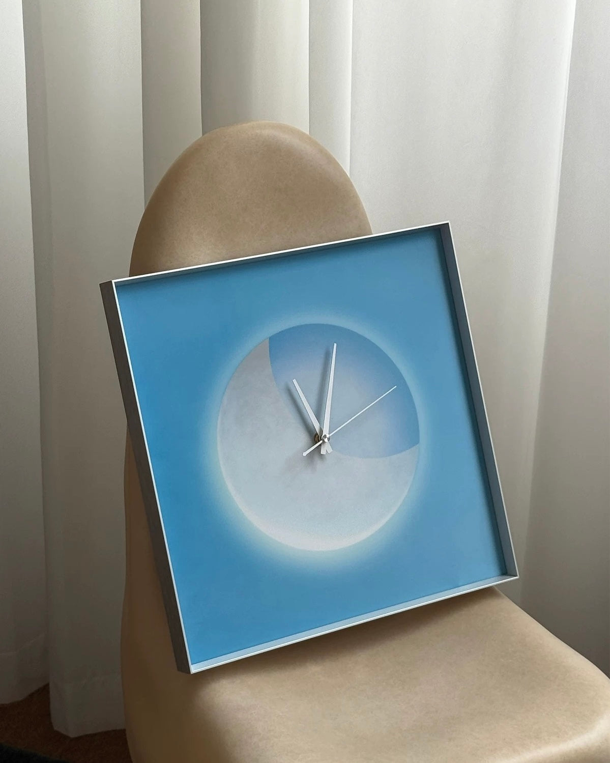 A square modern wall clock with a blue and white abstract design, resting on a beige upholstered chair in front of white curtains.