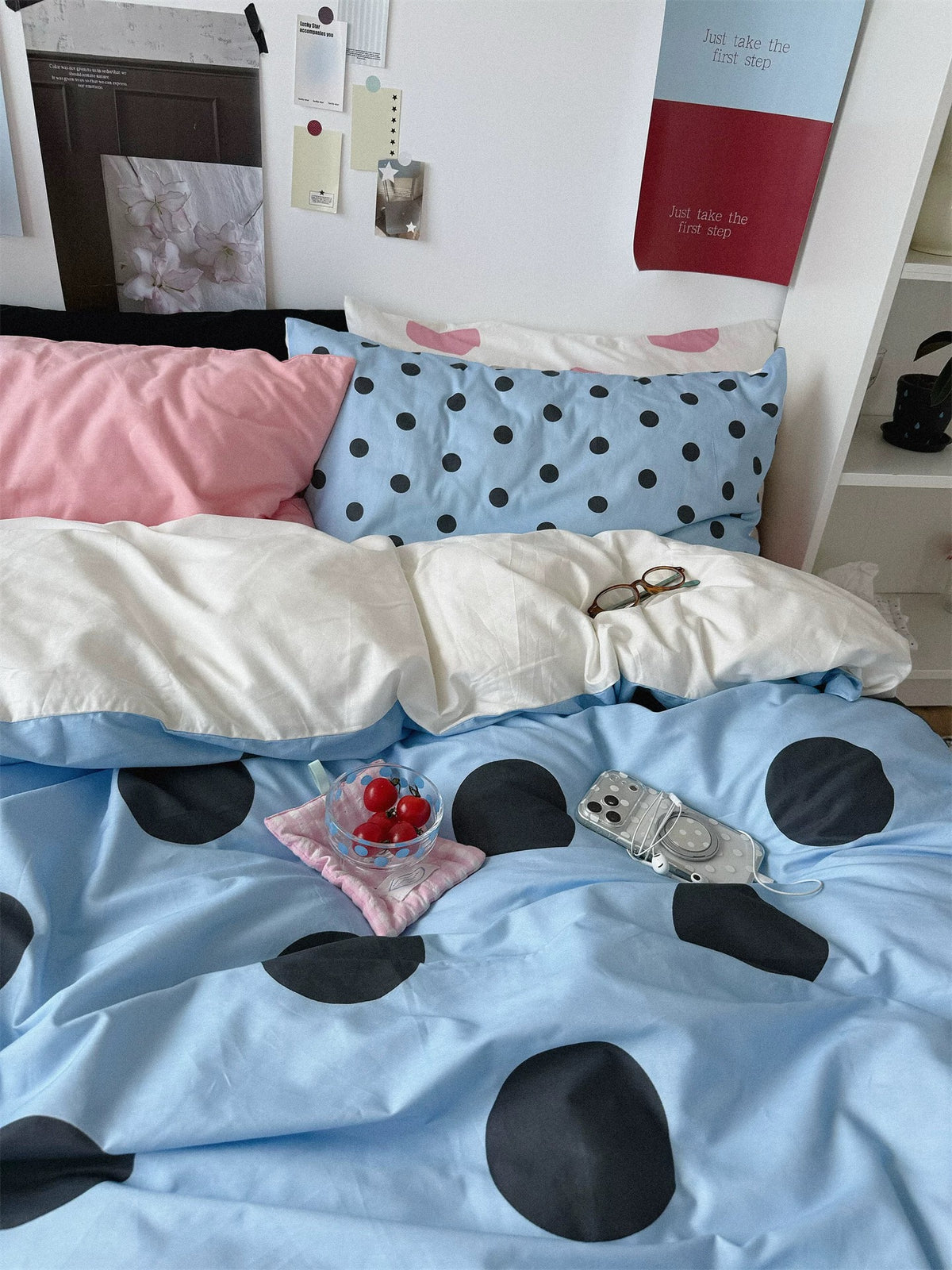 A blue duvet cover with large black dots lies below plain pink, small dotted blue and cream pillowcases.