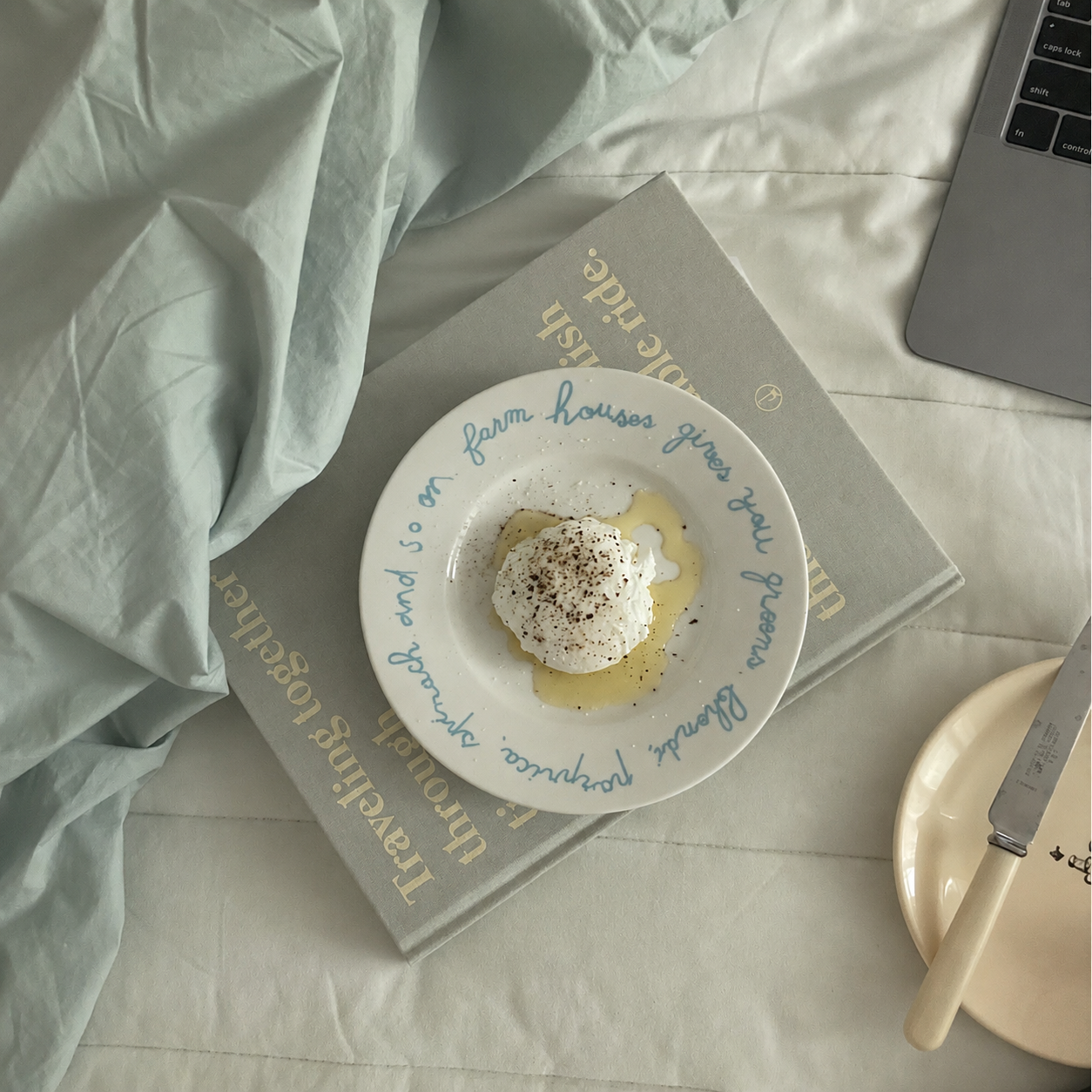 A white ceramic plate with blue cursive text, holding a poached egg with a drizzle of honey and black pepper, styled on top of a gray book with gold lettering on a white bed alongside a laptop and a cream-colored plate with a knife.