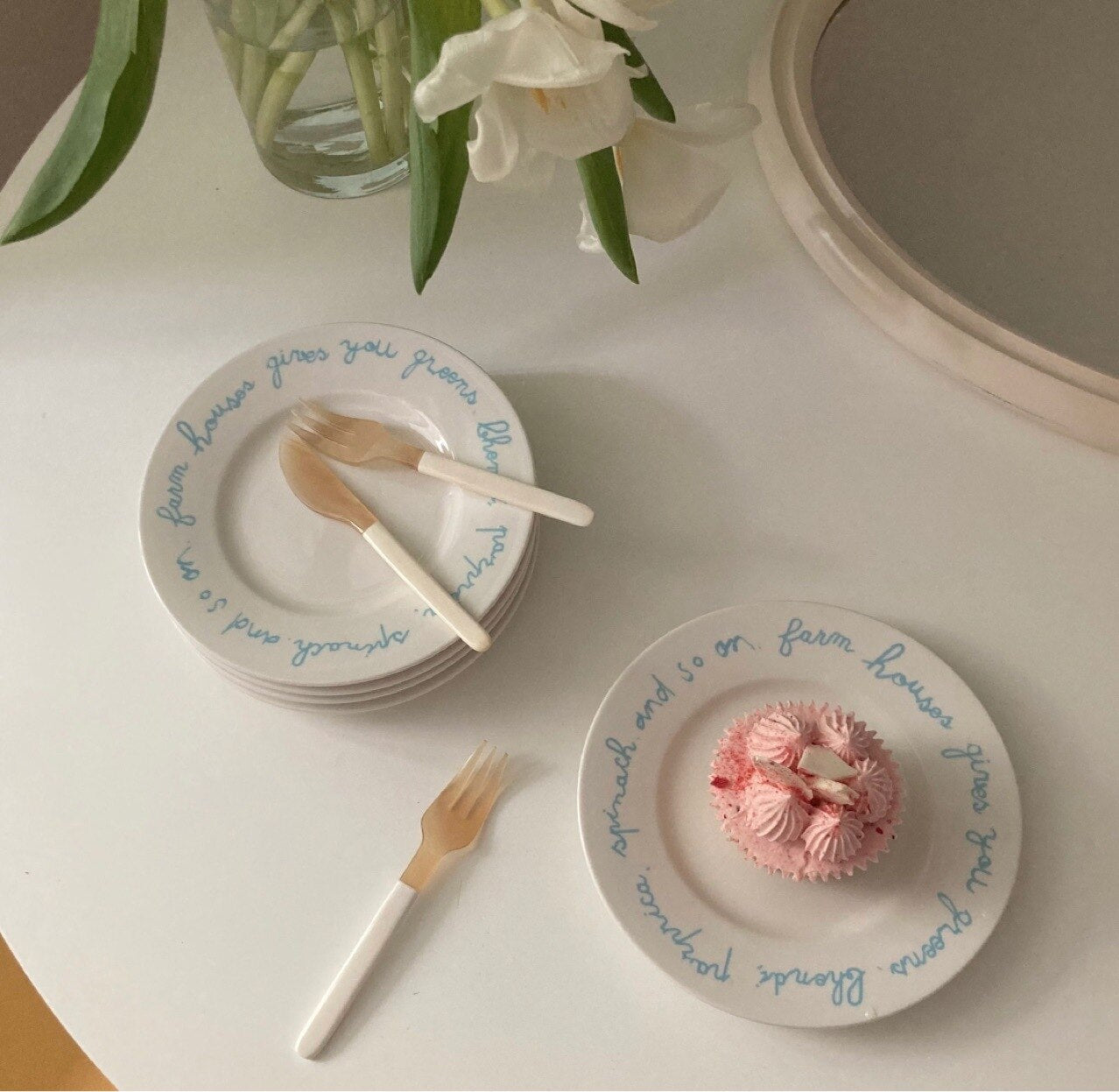 White ceramic plates with blue cursive text, accompanied by white-handled utensils, arranged on a white table with flowers and a pink cupcake in the background.