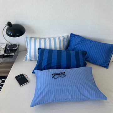 Three blue and white striped and pinstriped pillows arranged on a bed next to a black desk lamp and notebook.
