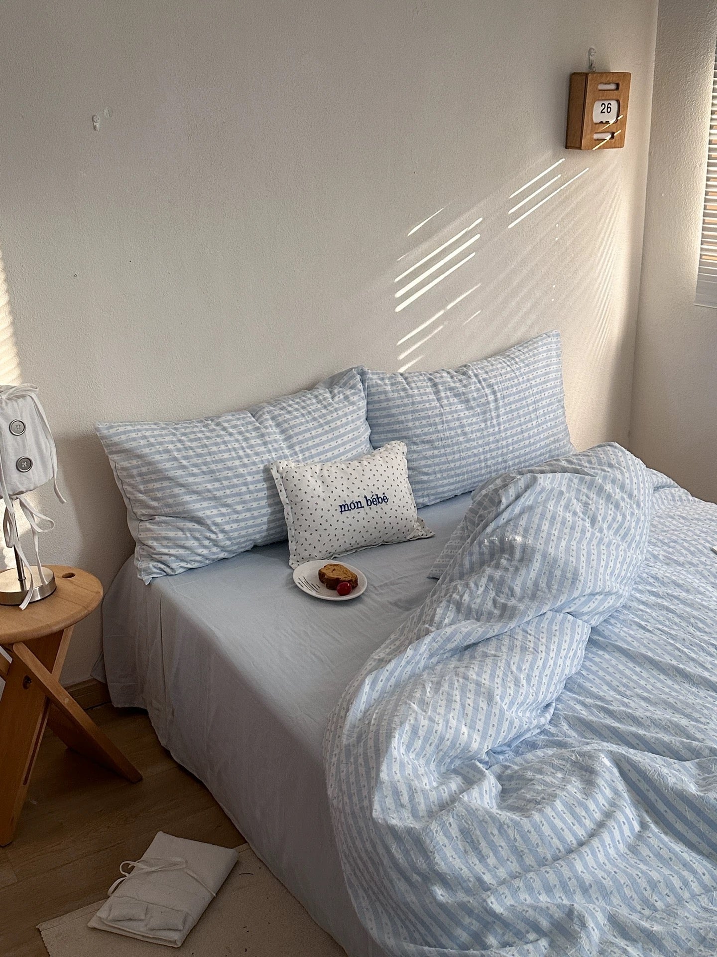 A neatly made bed in a sunlit bedroom with light blue and white striped bedding, matching pillows, a small white accent pillow, and a wooden side table beside it.