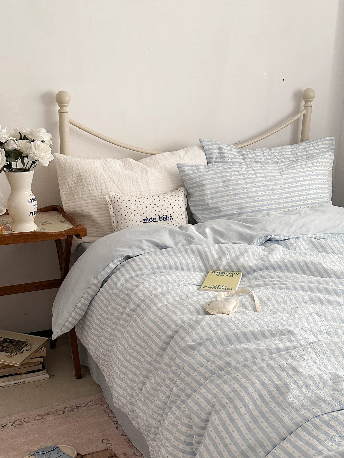 A neatly made bed with light blue and white striped bedding, including a duvet cover and matching pillowcases, styled in a cozy bedroom setting with a wooden side table, white flowers, and a small decorative pillow.
