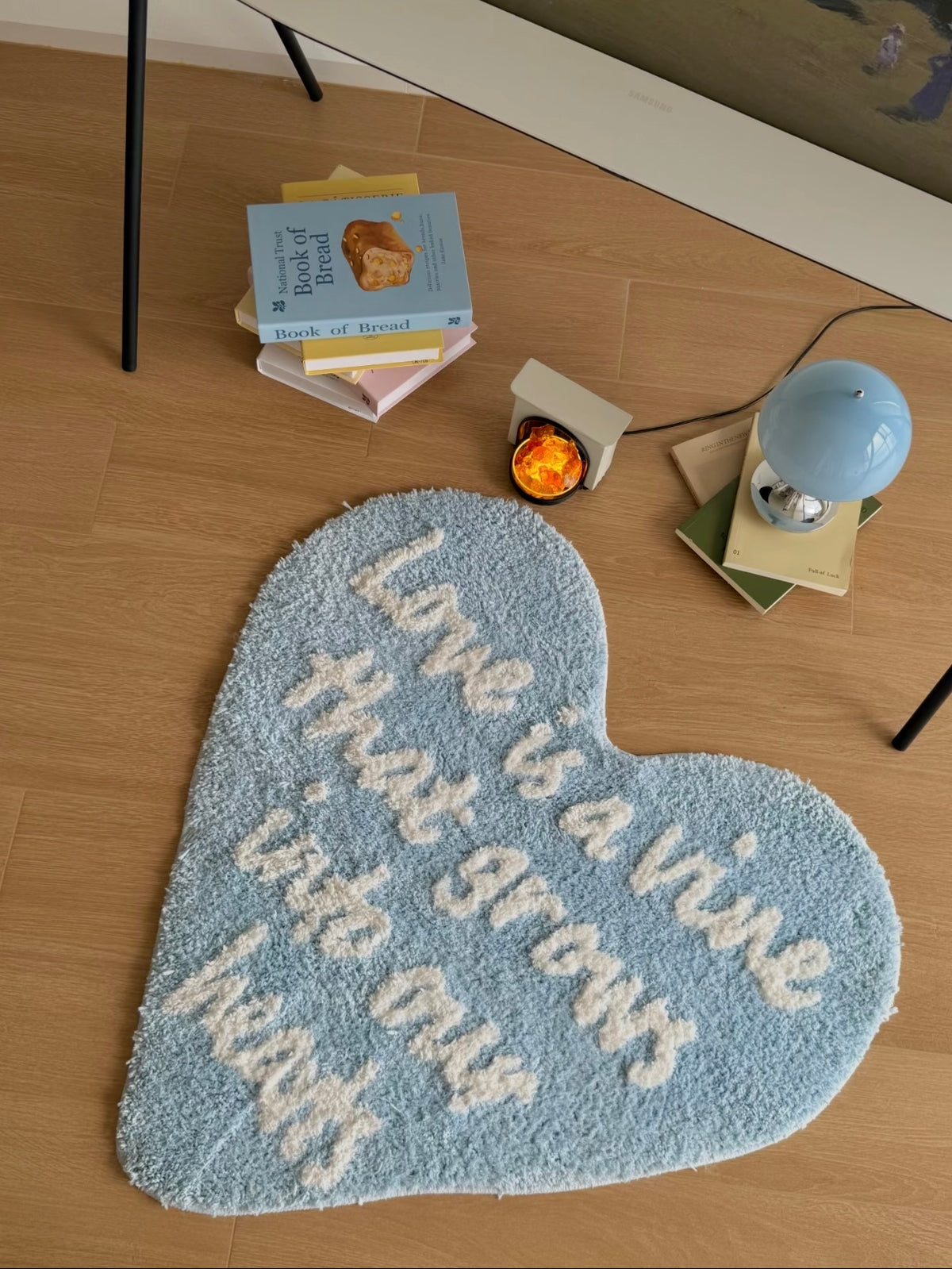 Blue heart rug with white lettering lies on a wooden floor, with books and a small blue lamp nearby.