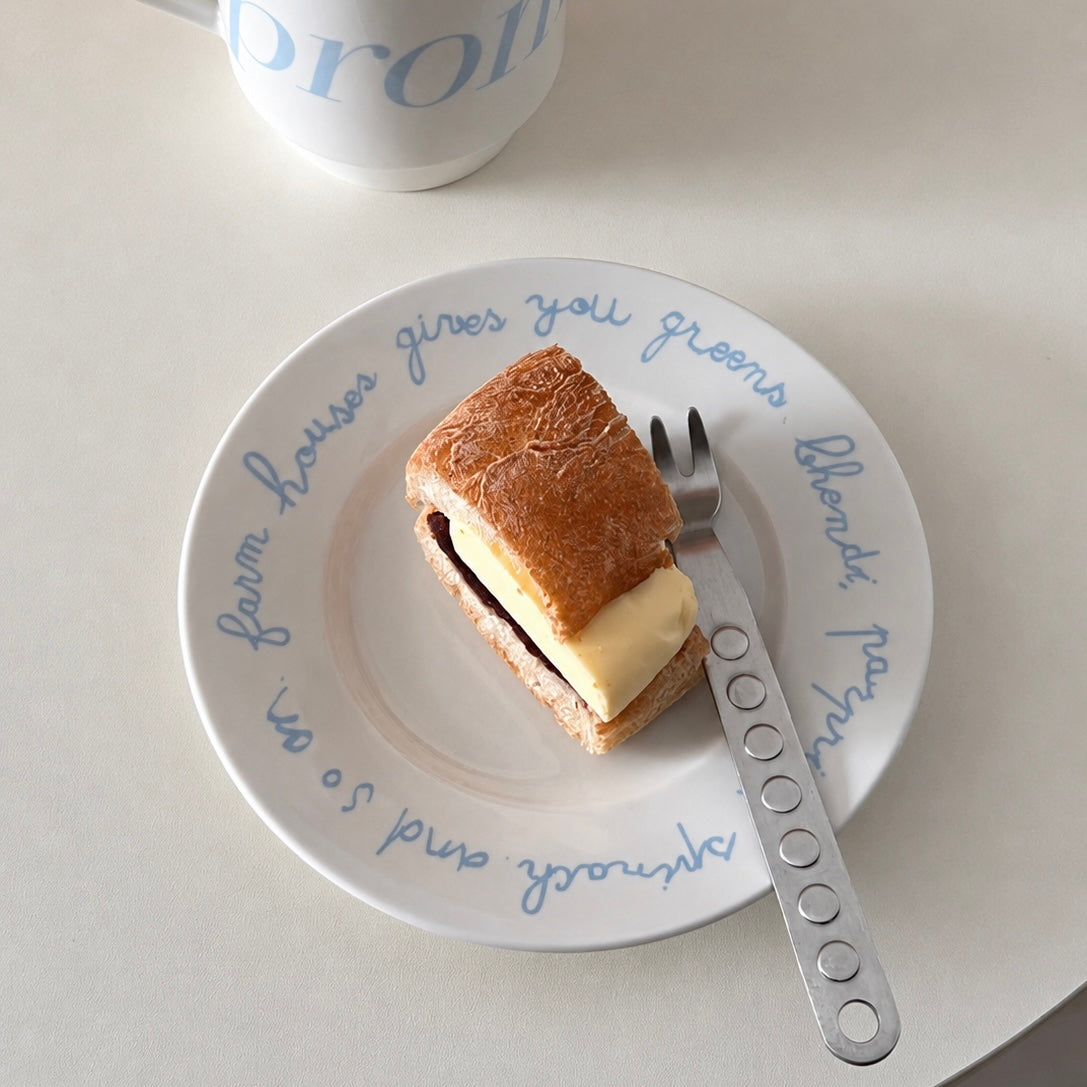 A slice of golden-brown bread with a thick pale yellow filling sits on a white plate with blue cursive text, next to a silver fork on a light beige tabletop.