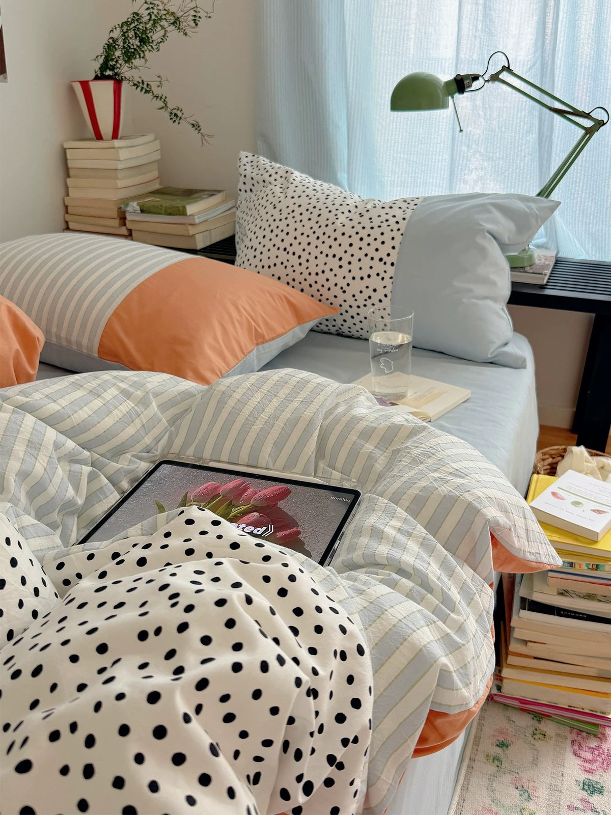Blue and yellow bedding with narrow stripes lies beneath dotted and peach-panel pillows beside a green desk lamp.