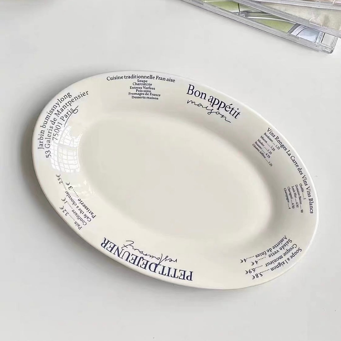 A breakfast plate from Petit Défonder featuring a golden-brown bagel sandwich with cheese and greens, served with two halves of a hard-boiled egg with runny yolks. The white plate has red French text and illustrations, set on a patterned beige tablecloth next to a glass of juice and a knife.