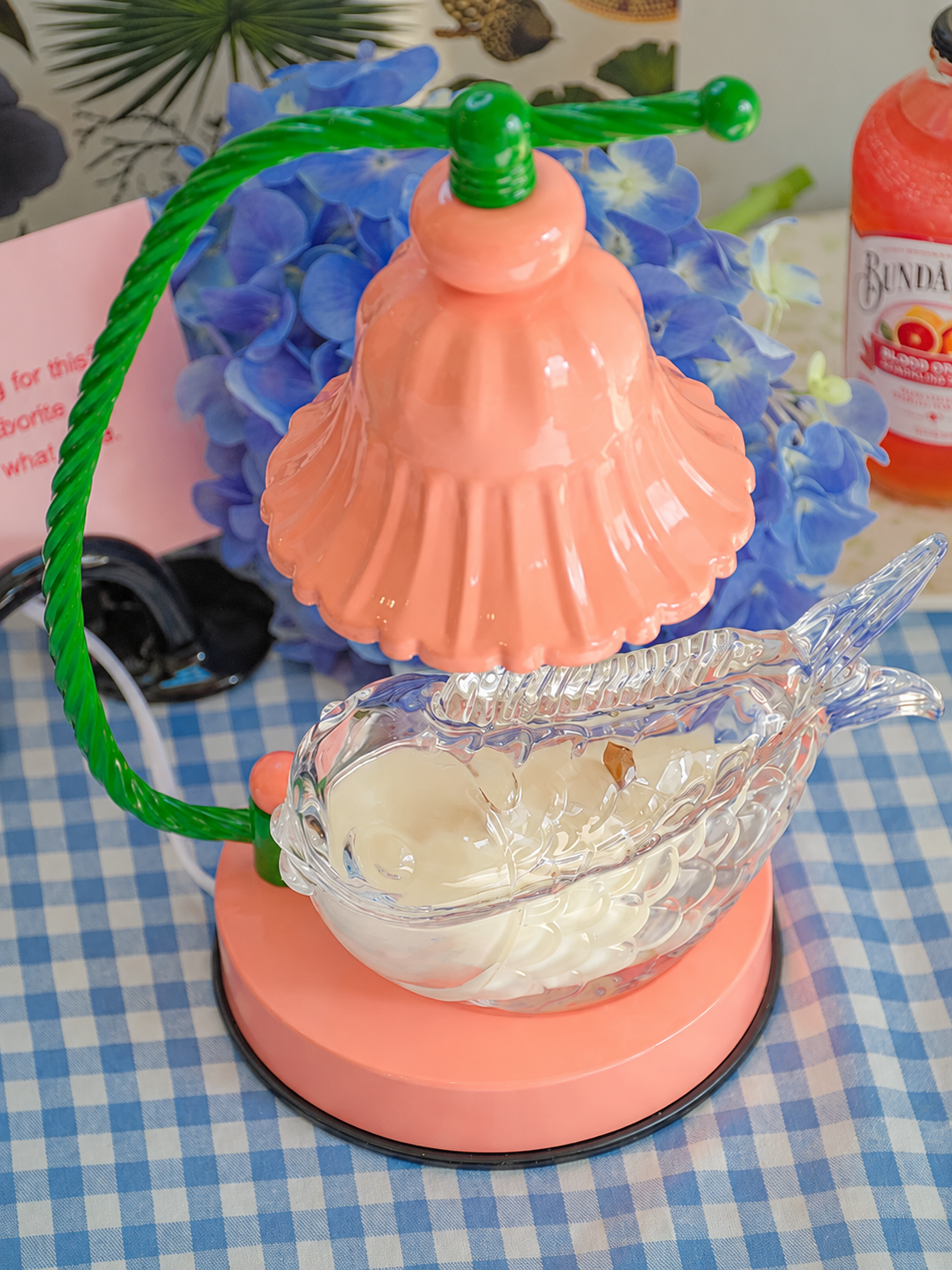 A decorative table lamp with a coral-pink bell-shaped shade, a green twisted metal arm, and a clear fish-shaped glass bowl filled with white wax, set on a blue-and-white checkered surface.