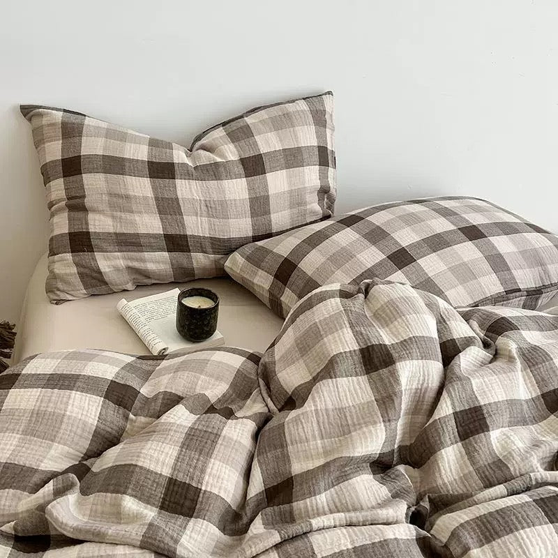 A cozy bed styled with a brown and beige plaid bedding set, including two pillowcases and a matching duvet, with a small black candle and an open book on a light wall.