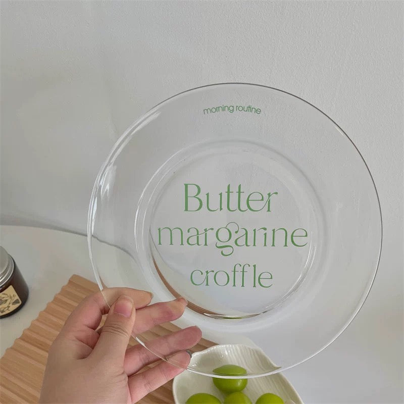 A hand holds a clear glass plate with green French text reading 'Butter margarine croffle' and small 'morning routine' text near the rim, against a light wall with a small bowl of green grapes in the background.