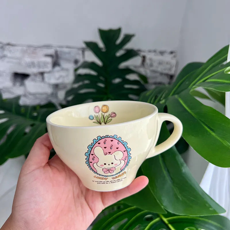 A hand holds a cream-colored ceramic teacup mug featuring a cute cartoon bunny character in a pink circle design, set against a background of green monstera leaves and a white brick wall.