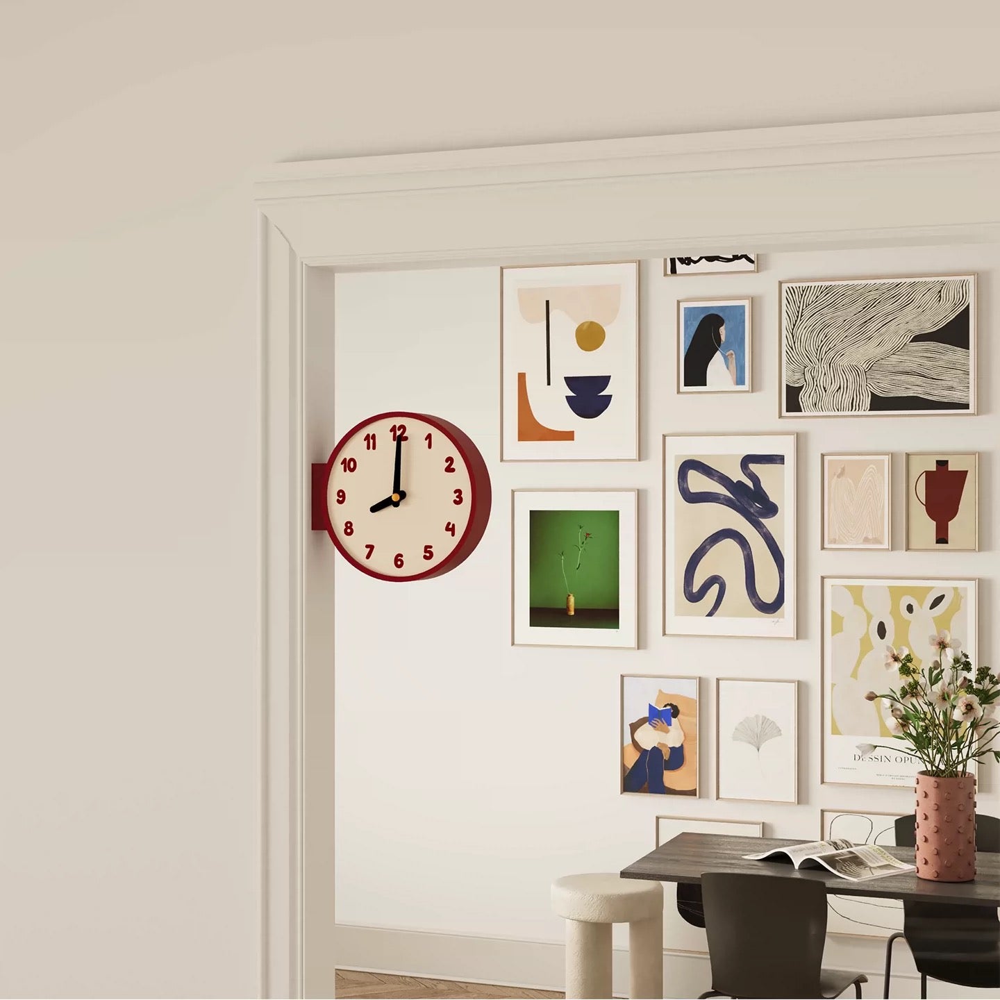 A styled interior scene showing a white wall with a gallery wall of framed abstract art prints, a round red clock, and part of a dining table with chairs and a vase of flowers.