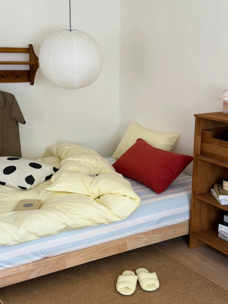 A cozy bedroom scene with a bed made up in pale yellow bedding, a blue-and-white striped sheet, a red pillow, a cream pillow, and a black-and-white polka dot accent pillow. A small round side table with a mug, book, and earbuds sits beside the bed.