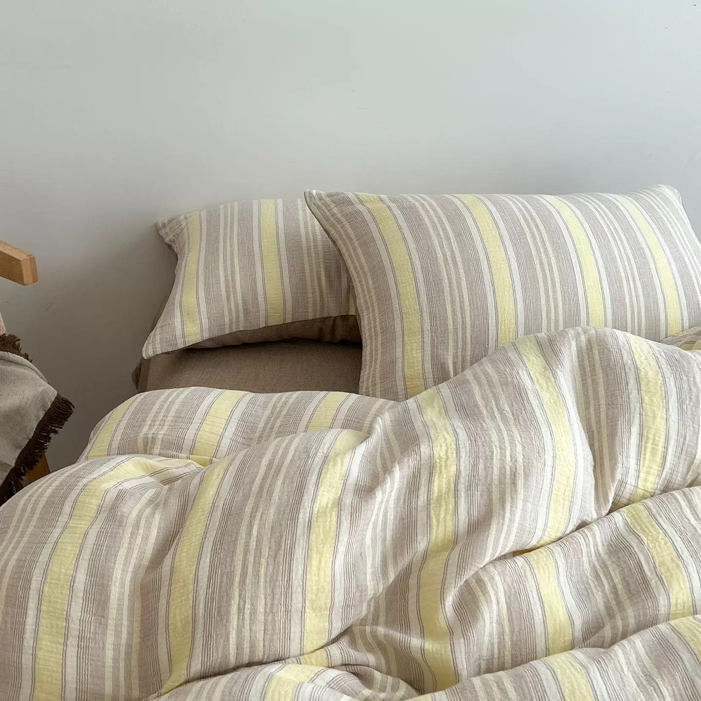 A striped bedding set with a duvet cover and two pillowcases in a soft beige, gray, and pale yellow stripe pattern, styled on a bed against a plain light gray wall.