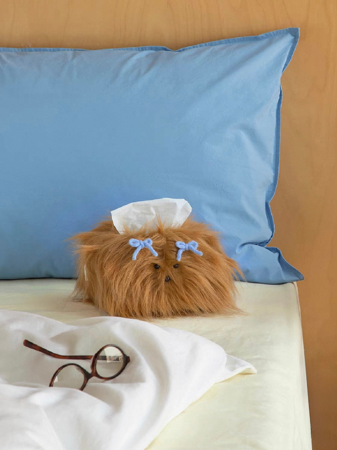 Caramel tissue holder on white bedding in front of a blue pillow, with a tissue rising above the bow clips.