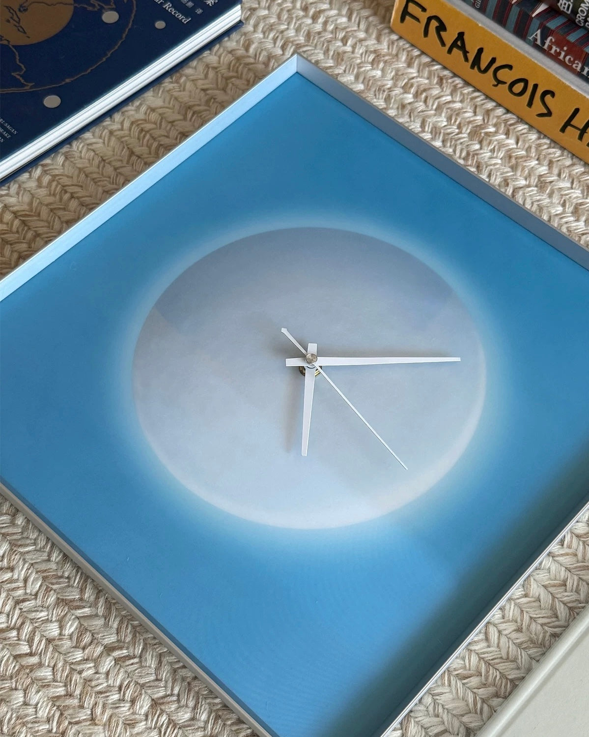 Square blue moon clock on a white wall above wooden shelving beside a light sofa.