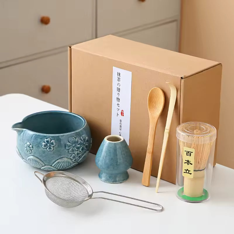 Tea-making set with a blue teapot, small vase, wooden spoon, and other tools on a white surface with a cardboard box in the background.