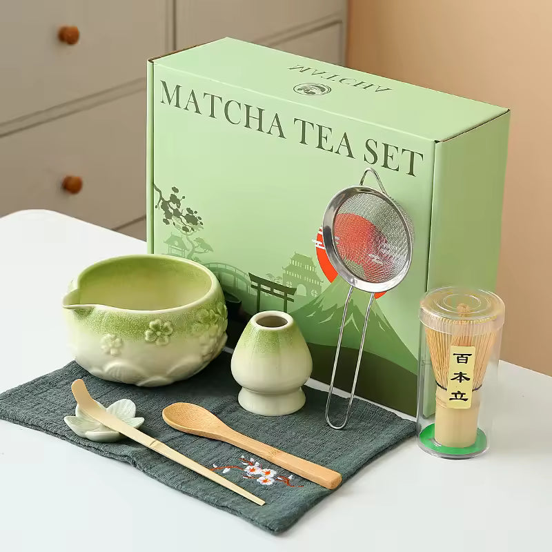 Matcha tea set with green box, ceramic bowl, whisk, and other tools on a white surface.