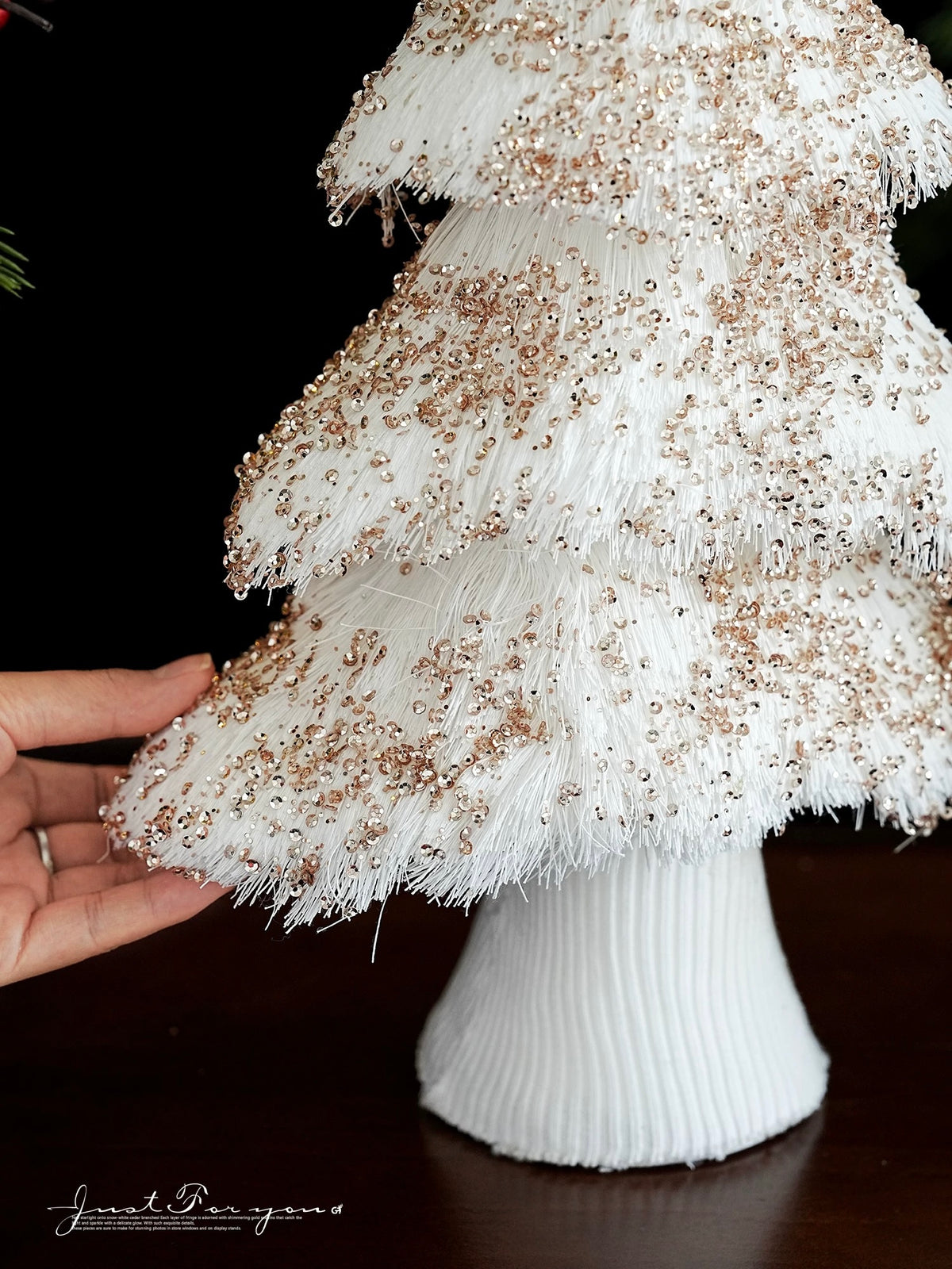 Champagne Christmas tree accents appear among white fringed branches while a hand lifts the edge of a tier.