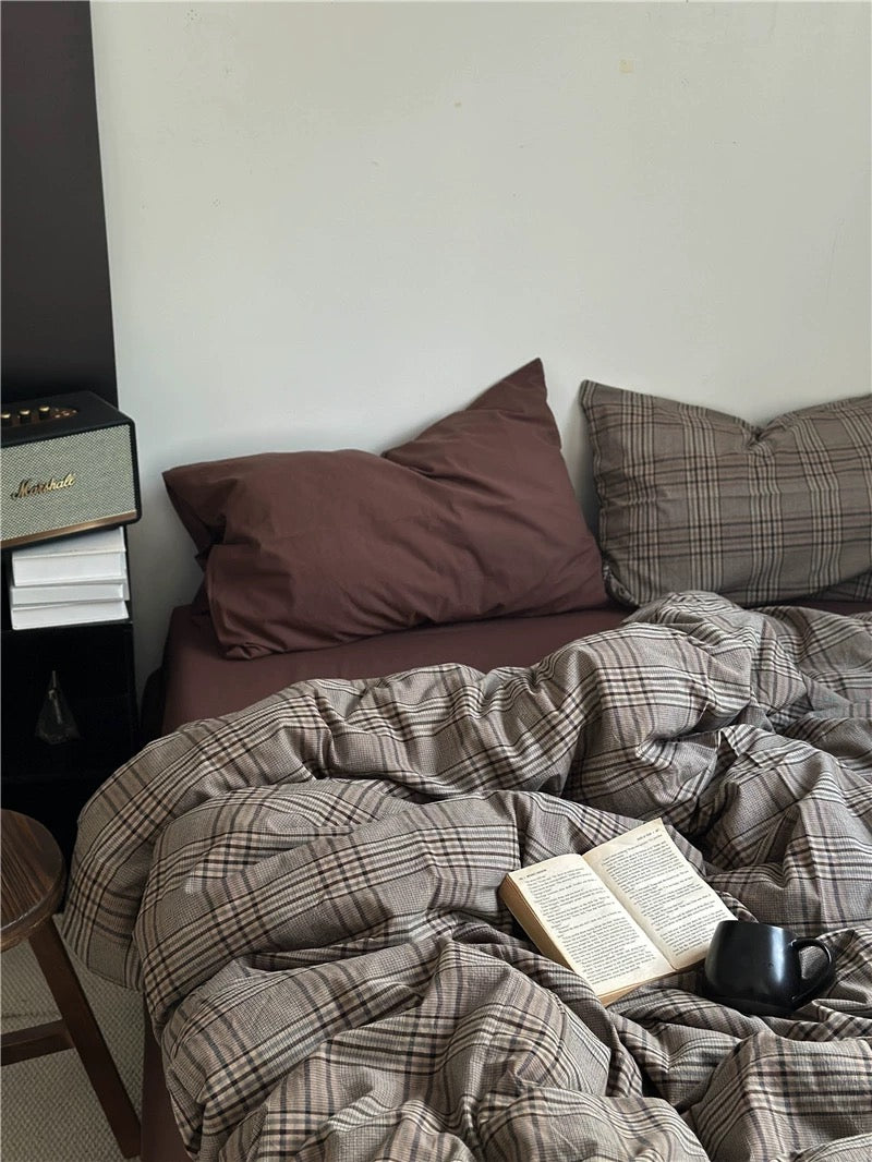 Bed with plaid comforter and pillows, book, and mug on a wooden nightstand.