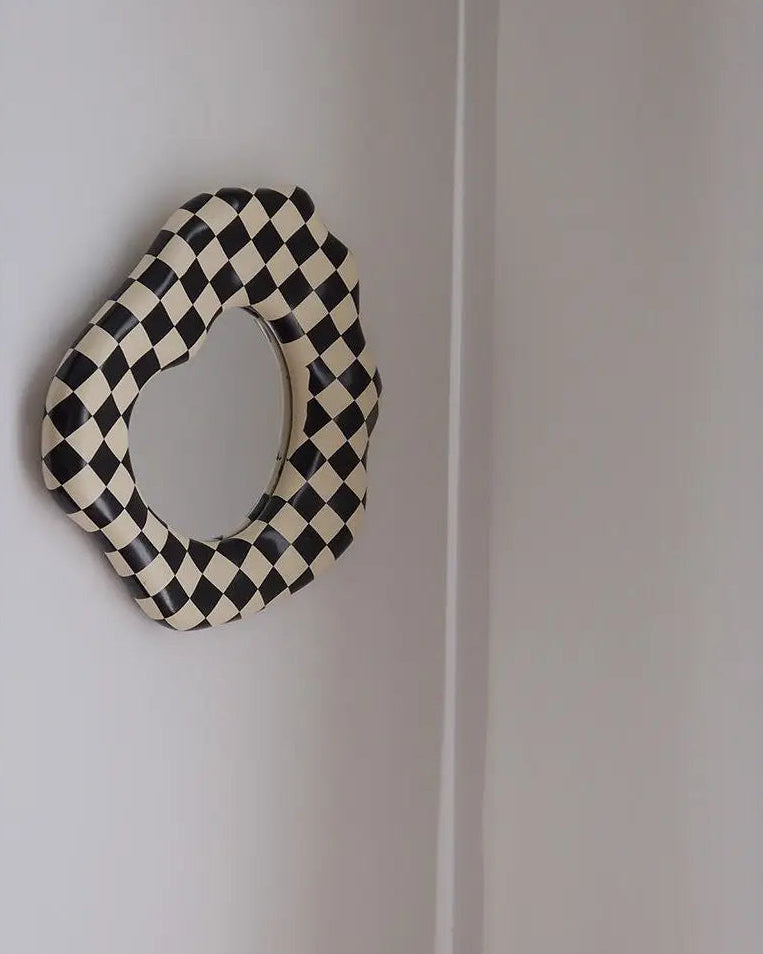An organic-shaped wall mirror with a black and cream checkerboard patterned frame, mounted on a plain white wall.