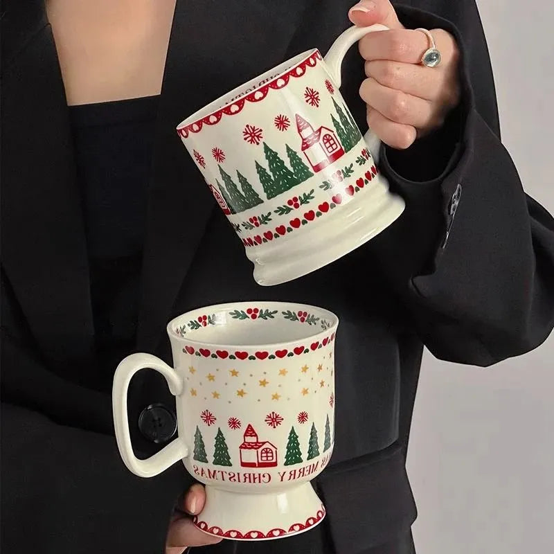 A person in a black blazer holds two ceramic mugs with a festive Christmas design. The mugs are cream-colored with red borders, green pine trees, red hearts, and a small red house with the text 'MERRY CHRISTMAS'.