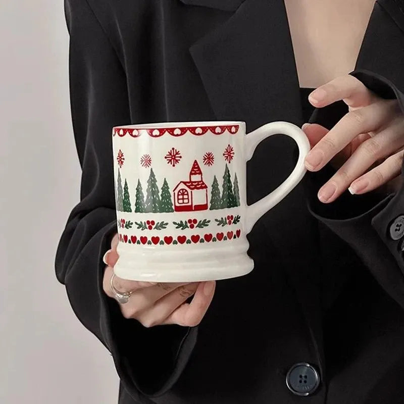 A person in a black blazer holds a white ceramic mug decorated with a red and green Christmas village pattern featuring a small house, trees, and snowflakes.