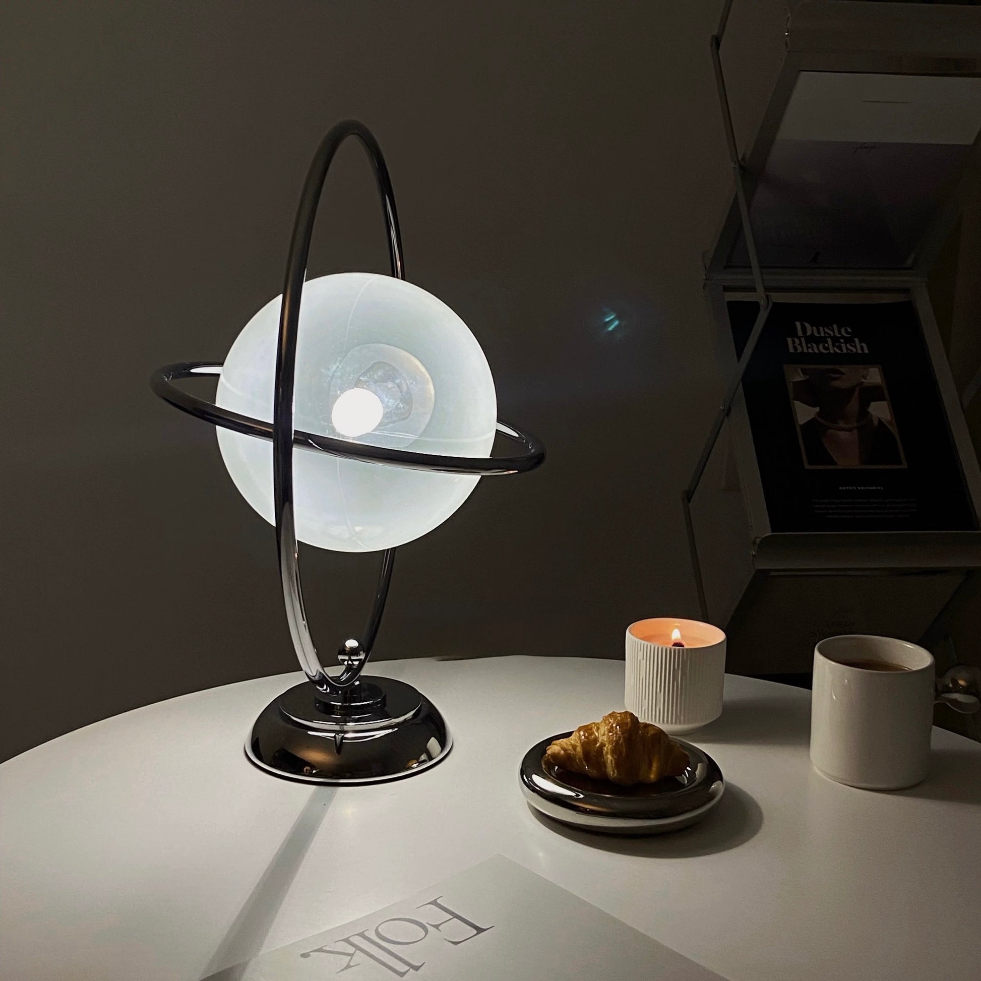 A modern table lamp with a frosted white globe and a black metal ring stand, placed on a white tabletop in a dimly lit room. A small candle, a ceramic cup, and a plate with a pastry are nearby, with a framed print and shelving in the background.