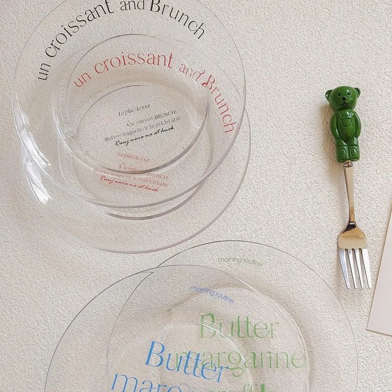 Two clear glass plates with printed French text and a small green bear-shaped fork on a textured cream surface.