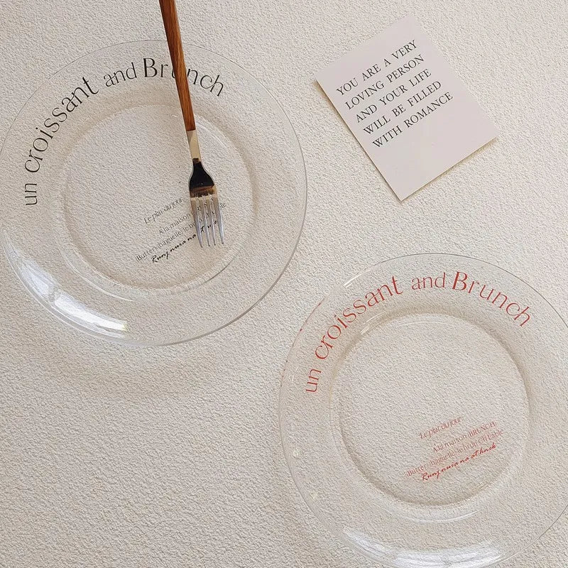 Two clear glass plates with printed French text, one in black and one in red, arranged on a textured cream background with a small card and a fork.