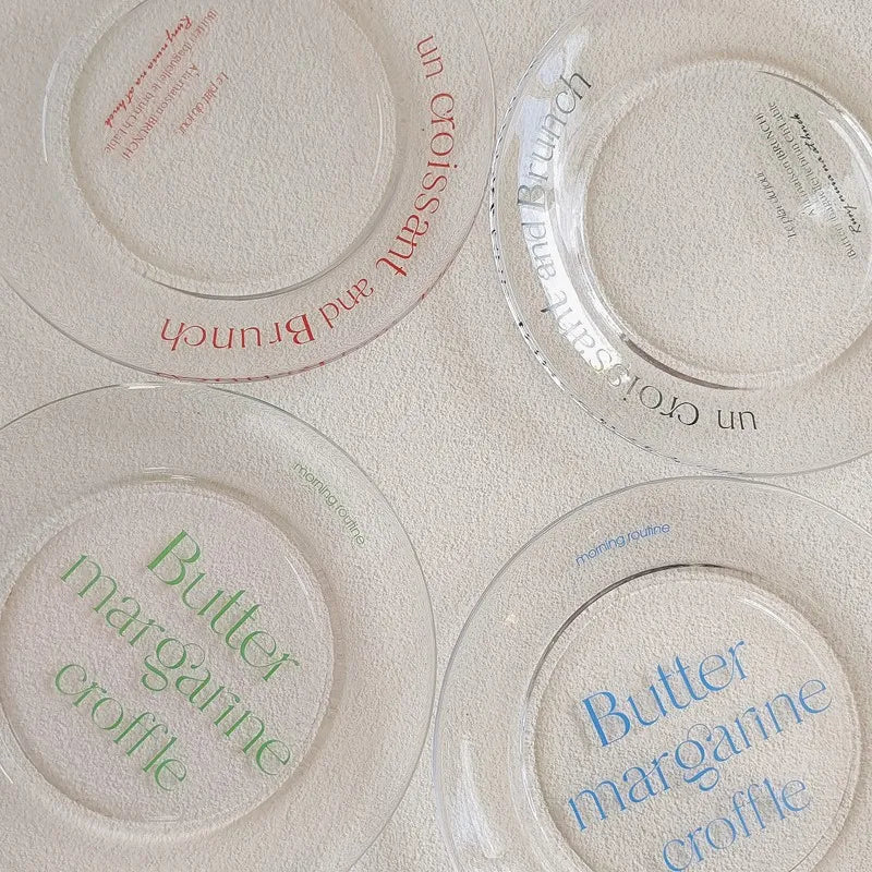 Four clear glass plates with French text printed in red, black, green, and blue, arranged on a textured off-white surface.